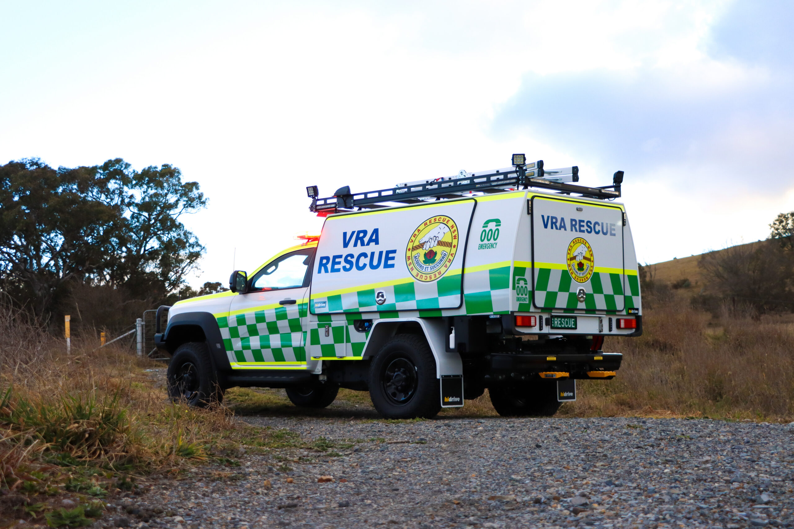 White and green VRA Rescue emergency response vehicle with flashing lights parked on a gravel track in a rural bushland setting. The four-wheel-drive utility features a custom Hidrive service body, roof-mounted equipment racks, and prominent VRA Rescue and Triple Zero Emergency signage. Dry grass, scattered trees, and rolling hills surround the vehicle beneath a cloudy sky.