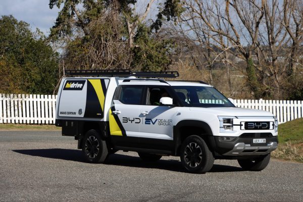 White BYD utility vehicle fitted with a Hidrive service body and roof rack, branded for BYD EV Dealer Group, parked on a paved roadside with a white picket fence and leafless trees in the background.