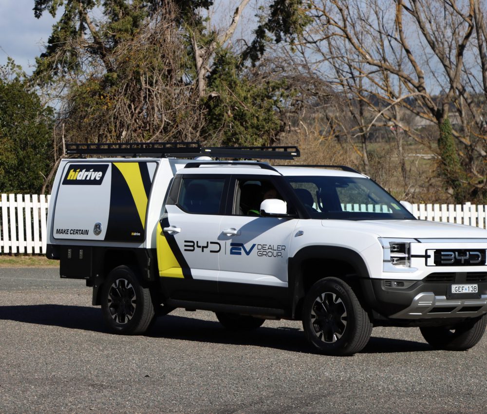 White BYD utility vehicle fitted with a Hidrive service body and roof rack, branded for BYD EV Dealer Group, parked on a paved roadside with a white picket fence and leafless trees in the background.