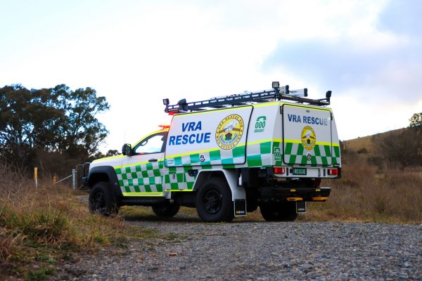 White and green VRA Rescue emergency response vehicle with flashing lights parked on a gravel track in a rural bushland setting. The four-wheel-drive utility features a custom Hidrive service body, roof-mounted equipment racks, and prominent VRA Rescue and Triple Zero Emergency signage. Dry grass, scattered trees, and rolling hills surround the vehicle beneath a cloudy sky.